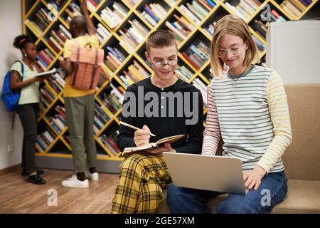 Portrait von zwei fröhlichen Studenten, die zusammen einen Laptop benutzen, während sie in der Universitätsbibliothek arbeiten, Platz kopieren Stockfoto