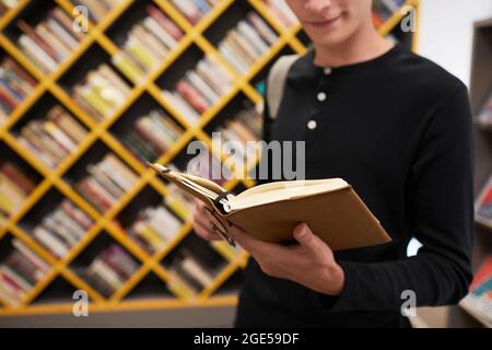 Ausgeschnittene Aufnahme eines jungen männlichen Studenten, der Buch hält, während er in der Schulbibliothek gegen Grafikregale steht und Platz zum Kopieren hat Stockfoto