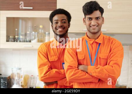 Portrait von glücklichen Klempnern, die sich die Arme kreuzen und die Kamera lächeln, wenn sie nach der Befestigung der Spülrohre in der Küche des Kunden stehen Stockfoto
