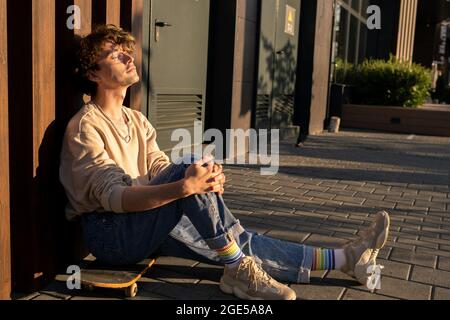 Junger entspannter Mann in Casualwear, der bei Sonnenuntergang auf dem Skateboard sitzt Stockfoto