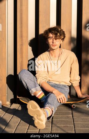 Ein hübscher Kerl in Jeans und Sweatshirt, der an der Wand auf dem Skateboard sitzt Stockfoto