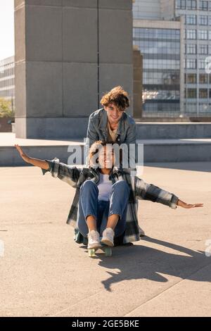 Fröhlicher Teenager, der seine afrikanische Freundin auf Skateboard schiebt, während beide Spaß haben Stockfoto