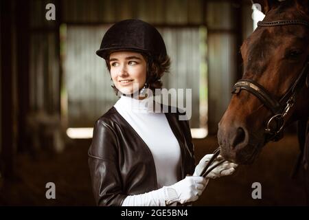 Nettes Mädchen Jockey, neben schönen braunen Pferd schießen Nahaufnahme. Schönes junges Mädchen steht neben Pferd im Stall. Sie trägt ein schönes weißes Kleid. Ranch. Reiten. Hippodrome Stockfoto