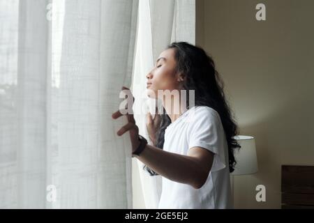Ruhiger, hübscher junger Mann mit langen lockigen Haaren, die Vorhänge öffnen und die ersten Sonnenstrahlen genießen Stockfoto