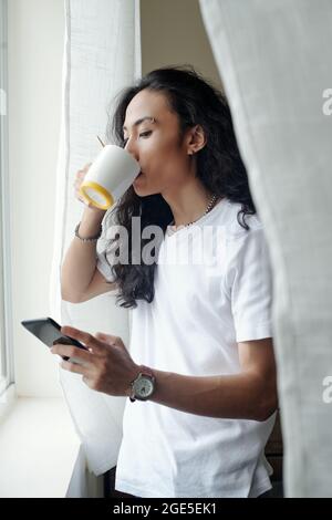 Hübscher junger Mann mit Augenbrauenpiercing, der Morgenkaffee trinkt, Nachrichten liest und Benachrichtigungen auf dem Smartphone überprüft Stockfoto