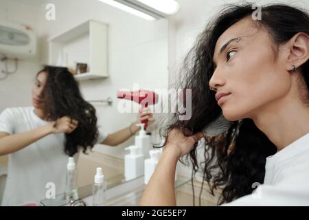 Schöner junger Mann, der seine langen lockigen Haare vor den Badezimmerspiegel bläst Stockfoto
