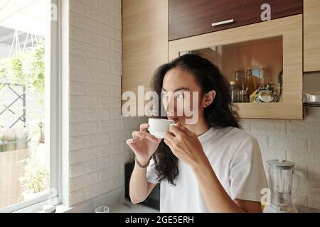 Porträt eines hübschen jungen Mannes, der eine Tasse Morgenkaffee trinkt und durch das Fenster schaut Stockfoto