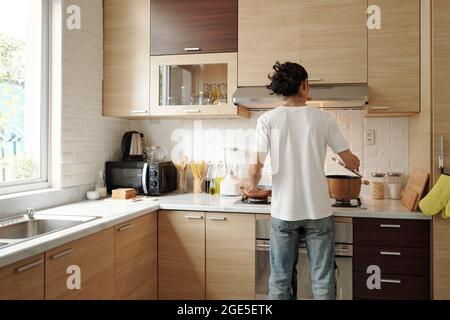 Junger Mann in Jeans und weißem Hemd kocht Abendessen an der Küchentheke, Blick von hinten Stockfoto