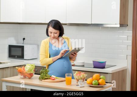 Schwangere Frau mit digitalen Tablet in der Nähe von Lebensmitteln auf Küchentisch Stockfoto