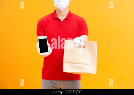 Junger gebender Mann mit medizinischen Handschuhen und Schutzmaske, der Smartphone mit Kopierraum und Einkaufstaschen in den Händen hält. Stockfoto