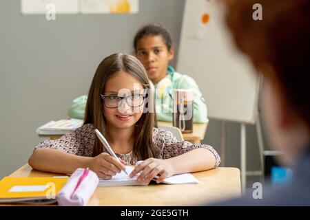 Glückliche Schülerin in Brillen Blick auf Lehrer, während Notizen in Copybook in der Lektion Stockfoto