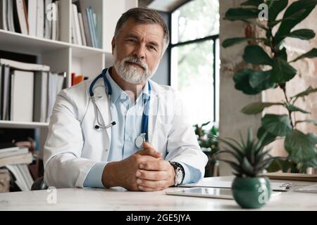 Porträt eines hübschen reifen Arztes, der im modernen Büro am Schreibtisch sitzt. Stockfoto