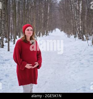 Eine glückliche Schwangere in einem Strickpullover und einem Hut spaziert im Park, einem Winterwald mit Bäumen im Schnee Stockfoto