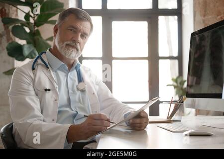 Porträt eines gut aussehenden, reifen Arztes, der im modernen Büro am Schreibtisch sitzt, sich Notizen macht oder die Krankenkarte des Kunden ausfüllt oder Medikamente verordnet. Medi Stockfoto