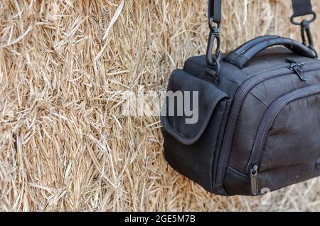 Nahaufnahme Schwarze Kameratasche, die an einer Wand aus trockenem Stroh hängt. Eine kleine wasserdichte Kameratasche. Lager von rechteckigen Heuballen. Stockfoto