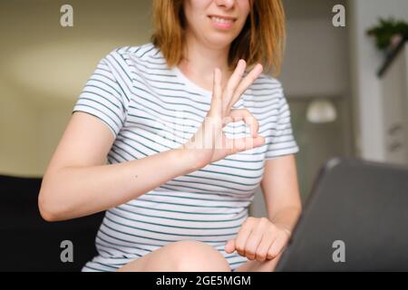 Lächelnde junge Frau mit Laptop für Online-Gespräche mit Hochschulen oder Freunden und zeigt Zeichen OK. Freiberuflicher Mitarbeiter, der zu Hause arbeitet. Stockfoto