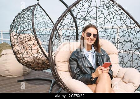 Porträt einer ziemlich lächelnden jungen Frau, die mit dem Smartphone in den Händen auf einem Stuhl sitzt und die Kamera anschaut Stockfoto