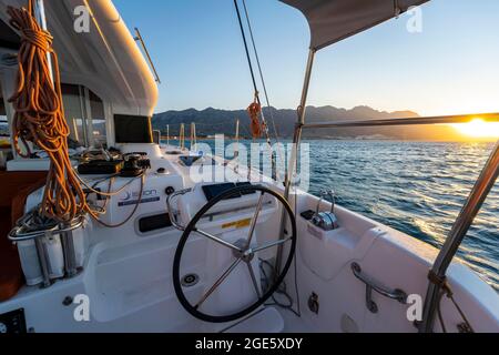 Lenkrad im Cockpit auf Deck eines Segel-Katamarans, Seile beim Segeln, Sonnenuntergang, Segeltörn, Dodekanes, Griechenland Stockfoto