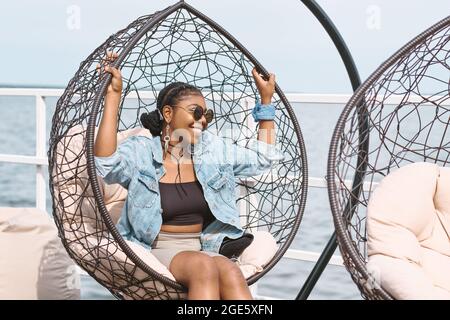 Fröhliche stilvolle junge Frau in Denim-Jacke, Sonnenbrille und langen Ohrringen sitzen in Hängesessel auf Outdoor-Geburtstagsfeier Stockfoto