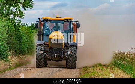 Ein gelber Traktor, der einen Anhänger schleppt, rast auf einer schmalen Feldbahn und kartiert zur Erntezeit Mais und wirft eine Staubwolke dahinter auf Stockfoto