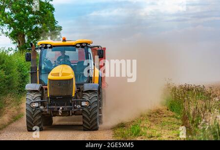 Ein gelber Traktor, der einen Anhänger schleppt, rast auf einer schmalen Feldbahn und kartiert zur Erntezeit Mais und wirft eine Staubwolke dahinter auf Stockfoto