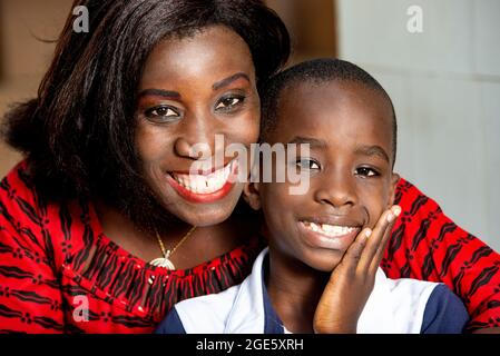 Schöne Erwachsene afrikanische Frau unter einem roten Lendenschurz und ihr Kind in weiß blauem T-Shirt machen ein schönes Lächeln zusammen Stockfoto