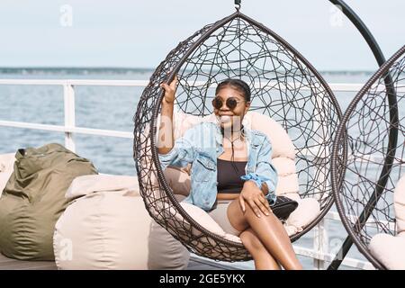 Portrait einer attraktiven lächelnden jungen Frau in Sonnenbrillen, die sich auf einem hängenden Stuhl auf der Party im Freien entspannt Stockfoto