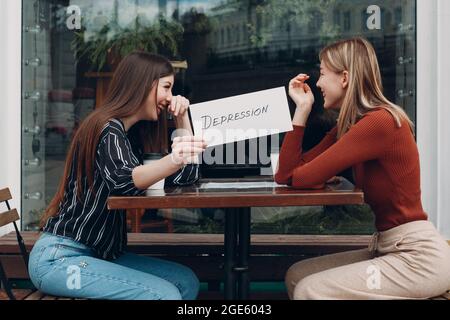 Konzept der versteckten Depression. Frau hält weißes Blatt Papier beschriftet Wort Depression in der Hand. Zwei Frauen im Straßencafé, die sich unterhalten und lächeln Stockfoto