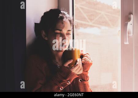Kalte Herbsttage - eine junge, multirassische Frau trinkt Kaffee in einer gemütlichen Fensterbank. Frau aus dem Nahen Osten oder Mischvolk trinkt am Wochenende gerne heißen Tee Stockfoto
