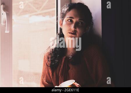 Kalte Herbsttage - eine junge, multirassische Frau trinkt Kaffee in einer gemütlichen Fensterbank. Frau aus dem Nahen Osten oder Mischvolk trinkt am Wochenende gerne heißen Tee Stockfoto