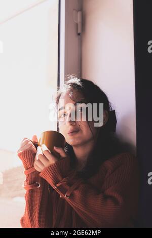 Kalte Herbsttage - eine junge, multirassische Frau trinkt Kaffee in einer gemütlichen Fensterbank. Frau aus dem Nahen Osten oder Mischvolk trinkt am Wochenende gerne heißen Tee Stockfoto