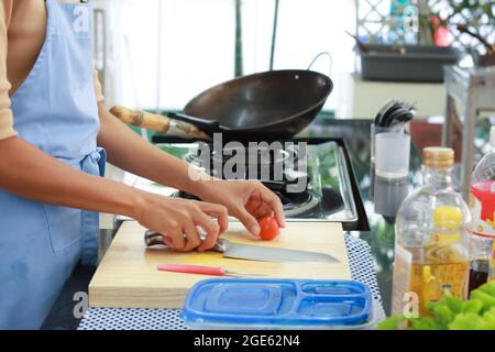 Körperteil einer asiatischen Frau mit blauer Schürze in der Küche. Hand halten Messer auf Schneidebrett zu schneiden Tomaten. Die Küche ist im asiatischen Stil mit ga Stockfoto