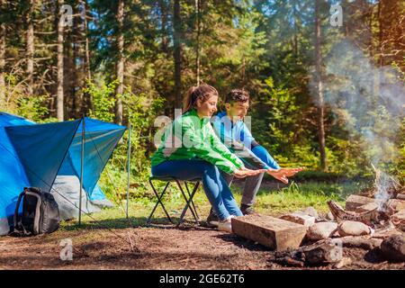 Junge Paare von Reisenden genießen Lagerfeuer im Sommerwald. Touristen sitzen am Zelt wärmende Hände. Mann und Frau entspannen Camping im Wald Stockfoto