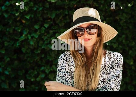 Nahaufnahme einer lächelnden jungen Frau mit Strohhut und Sonnenbrille, die draußen auf dem grünen Blatt-Hintergrund steht. Stockfoto