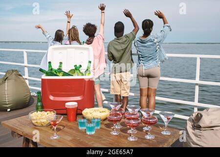 Tisch mit Kühlbox voller Bier, Snacks und Cocktal Drinks auf dem Tisch, junge Leute winken und sehen das Wasser im Hintergrund Stockfoto