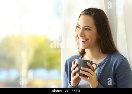 Eine fröhliche, nachdenkliche Frau schaut neben einem Fenster und hält zu Hause eine Kaffeetasse Stockfoto