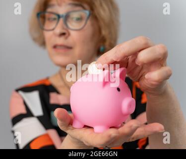 Eine ältere Frau mit Brille legt eine Münze in ein Sparschwein auf weißem Hintergrund Stockfoto