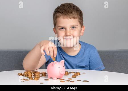 Boy lächelt und legt Münze in das Sparschwein. Das Kind investiert in die zukünftige Bildung. Stockfoto