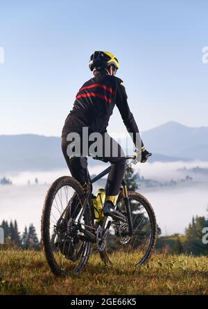 Rückansicht eines jungen Mannes im Radanzug, der auf einem grasbewachsenen Hügel Fahrrad fährt. Männlicher Radfahrer, der während einer Radtour den Blick auf die majestätischen Berge genießt. Konzept von Sport und Radfahren. Vertikales Bild. Stockfoto