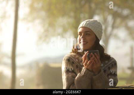 Eine fröhliche Frau am Morgen, die Kaffee trinkt und in einem nebligen Park sitzt und auf die Seite schaut Stockfoto