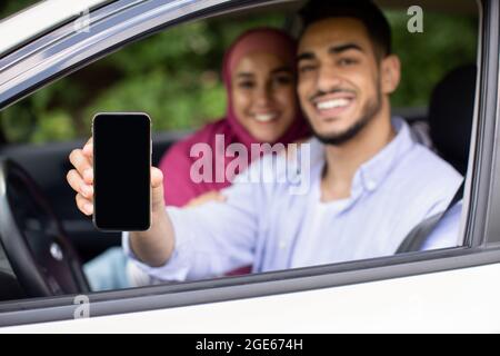 Glückliche Muslimische Ehegatten, Die Im Auto Sitzen Und Leeres Smartphone Mit Schwarzem Bildschirm Zeigen, Fröhlicher Mann Und Frau Aus Dem Nahen Osten In Hijab, Die New Empfehlen Stockfoto