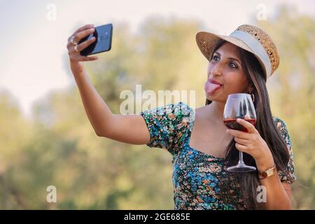 Lustige modische junge Frau mit Strohhut zeigt Zunge, macht Selfie mit Handy, hält Weinglas in den Händen, genießt Erholung und gute Summe Stockfoto