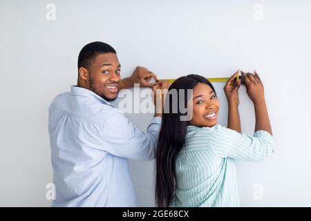 Glückliches schwarzes Paar, das die Wand mit Maßband misst und zusammen Reparaturen in einer neuen Wohnung durchführt. Junge afroamerikanische Ehegatten tun Heimverbesserung ein Stockfoto