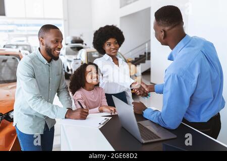 Es ist ein Deal. Positive schwarze Familie kauft neues Auto, schüttelt die Hände mit Verkäufer, Unterzeichnung Auto Kaufvertrag bei Händler. Fröhliche Afro-Kunden sh Stockfoto