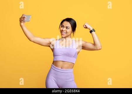 Selfie vom Trainer Blogger. Glücklich fit schwarze Frau in Sportbekleidung machen Foto auf dem Smartphone für Blog und zeigt ihren Bizeps, posiert auf gelben Backgrou Stockfoto