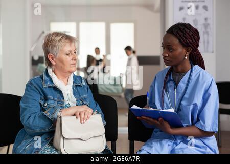 afroamerikanische Assistentin, die Krankheitssymptome mit älteren kranken älteren Patienten diskutiert und medizinische Behandlung im Wartezimmer des Krankenhauses auf die Zwischenablage schreibt. Support-Service im Gesundheitswesen Stockfoto