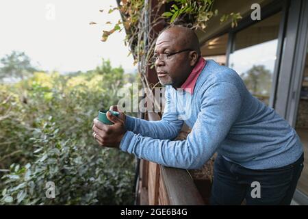 Nachdenklicher älterer afroamerikanischer Mann auf sonnigem Balkon, der eine Tasse Kaffee ausgießt Stockfoto