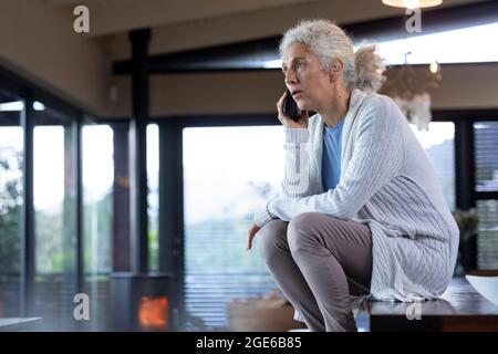 Ältere kaukasische Frau in der Küche, die auf dem Tisch sitzt und das Smartphone benutzt Stockfoto