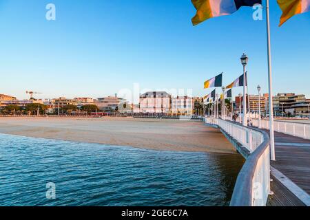 Arcachon (Südwestfrankreich): Der Thiers Pier, blaue, weiße und gelbe Flaggen der Stadt Arcachon, Gebäude entlang der Uferpromenade und leerer Strand Stockfoto