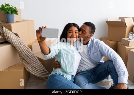 Glückliche schwarze Ehegatten, die in ihrer neuen Wohnung Selfie machen, den Tag des Umzugsens feiern und auf dem Boden in der Nähe von Kartons sitzen. Liebevoller Ehemann küsst mit Stockfoto